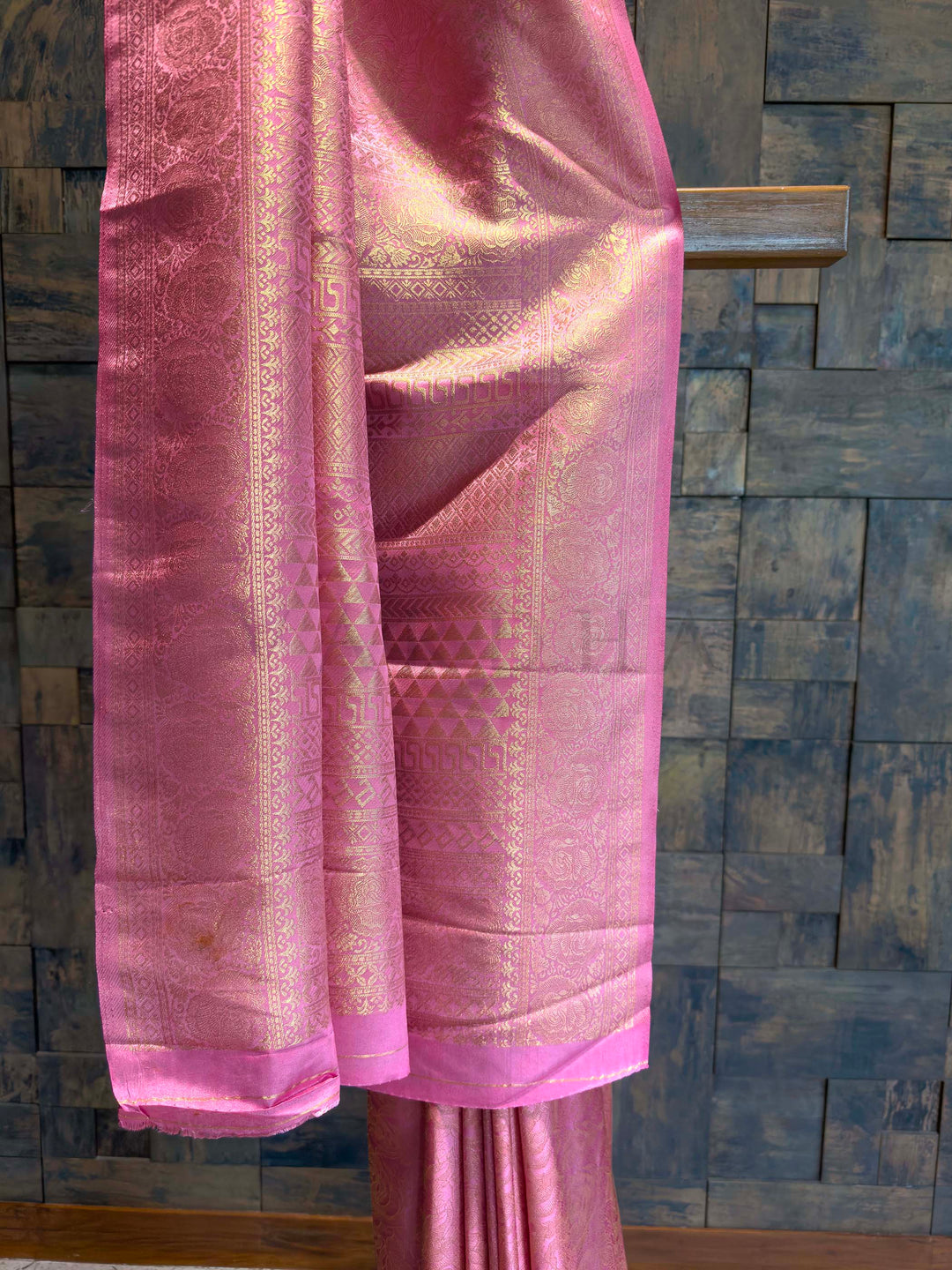 Pink fabric with intricate patterns draped over a textured wall. Floral and geometric golden zari patterns woven across rose pink Kanjivaram fabric
