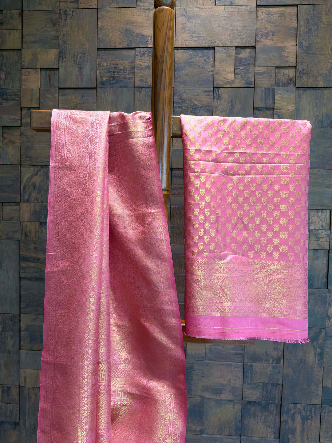Pink saree with a geometric pattern hanging on a wooden rod against a stone wall. Side profile showcasing border detailing and overall drape texture of the saree