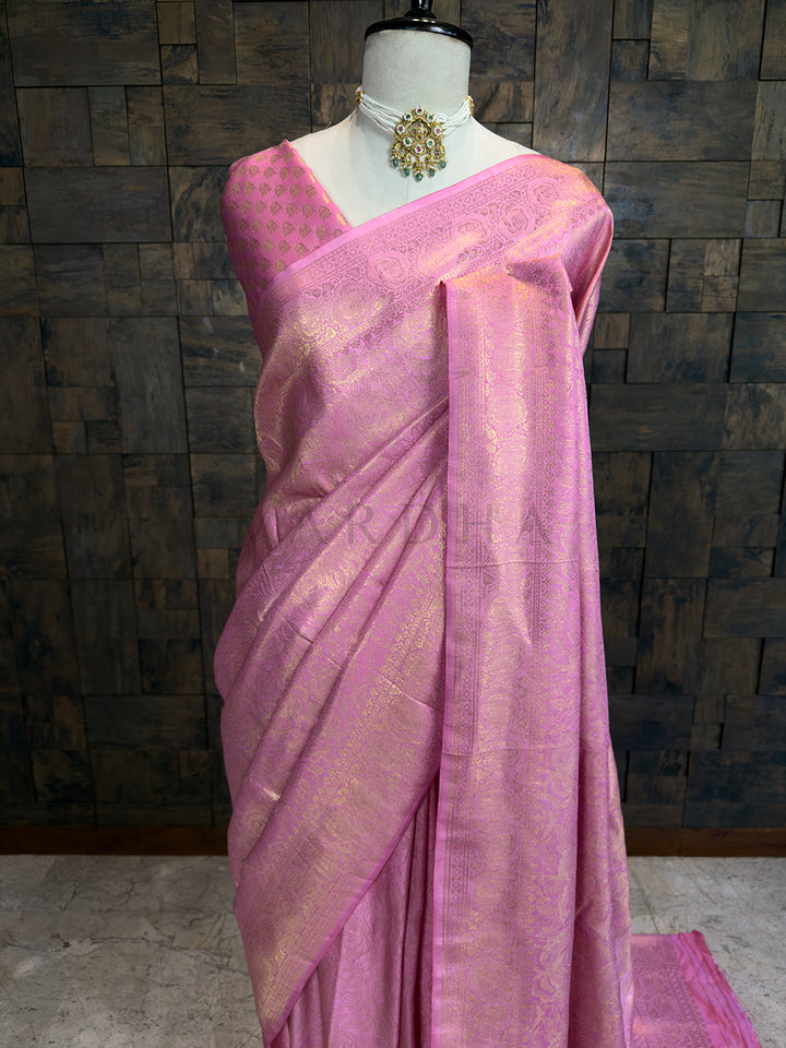Premium light pink Kanjivaram bridal saree by House of Vardha showcasing exquisite gold zari weaving and a rich traditional border. We offer Ready to wear, Pre drape service, Saree Add on, Custom Stitched Blouse, Fall and Edging, Gift Packaging at Complimentary Price! Pink saree with a textured pattern on a mannequin against a dark brick wall.