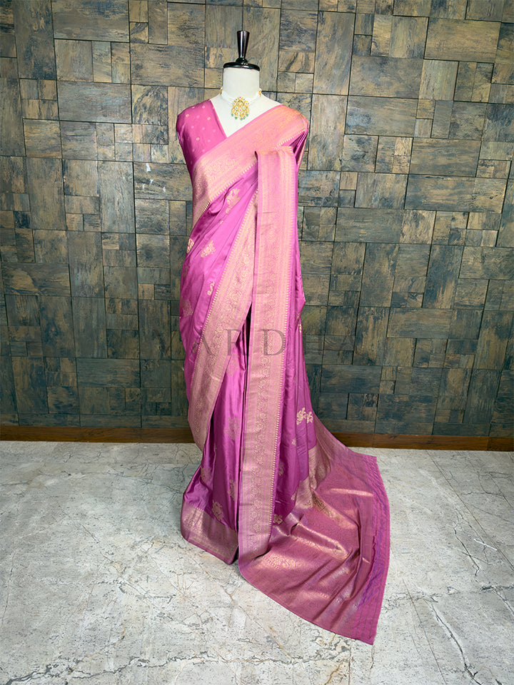 Pink saree with a mannequin against a textured wall background. House of Vardha Rose Pink Satin Saree featuring Antique Gold Zari Floral Motifs and a matching border, draped gracefully on a mannequin. We offer Ready to wear, Pre drape service, Saree Add on, Custom Stitched Blouse, Fall and Edging, Gift Packaging at Complimentary Price!