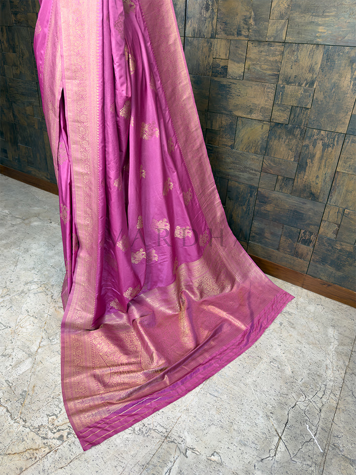 Pink saree with gold patterns draped over a textured wall. Luxurious Rose Pink Satin Saree from House of Vardha with heavy Antique Gold Zari embellishments, perfect for festive wear. We offer Ready to wear, Pre drape service, Saree Add on, Custom Stitched Blouse, Fall and Edging, Gift Packaging at Complimentary Price!