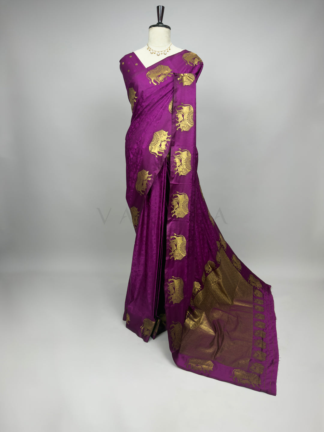 Purple saree with gold patterns on a mannequin against a gray background A full-length presentation of a Royal Magenta Satin Silk Saree elegantly draped on a mannequin against a neutral gray background. The saree features a vibrant, deep magenta body enhanced with a subtle self-jacquard floral texture, beautifully contrasted by large gold zari elephant motifs woven along the outer drape and shoulder. The heavy, richly woven gold pallu cascades fluidly onto the floor, creating a luxurious and regal festive l