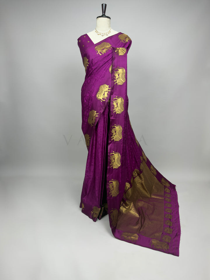 Purple saree with gold patterns on a mannequin against a gray background A full-length presentation of a Royal Magenta Satin Silk Saree elegantly draped on a mannequin against a neutral gray background. The saree features a vibrant, deep magenta body enhanced with a subtle self-jacquard floral texture, beautifully contrasted by large gold zari elephant motifs woven along the outer drape and shoulder. The heavy, richly woven gold pallu cascades fluidly onto the floor, creating a luxurious and regal festive l