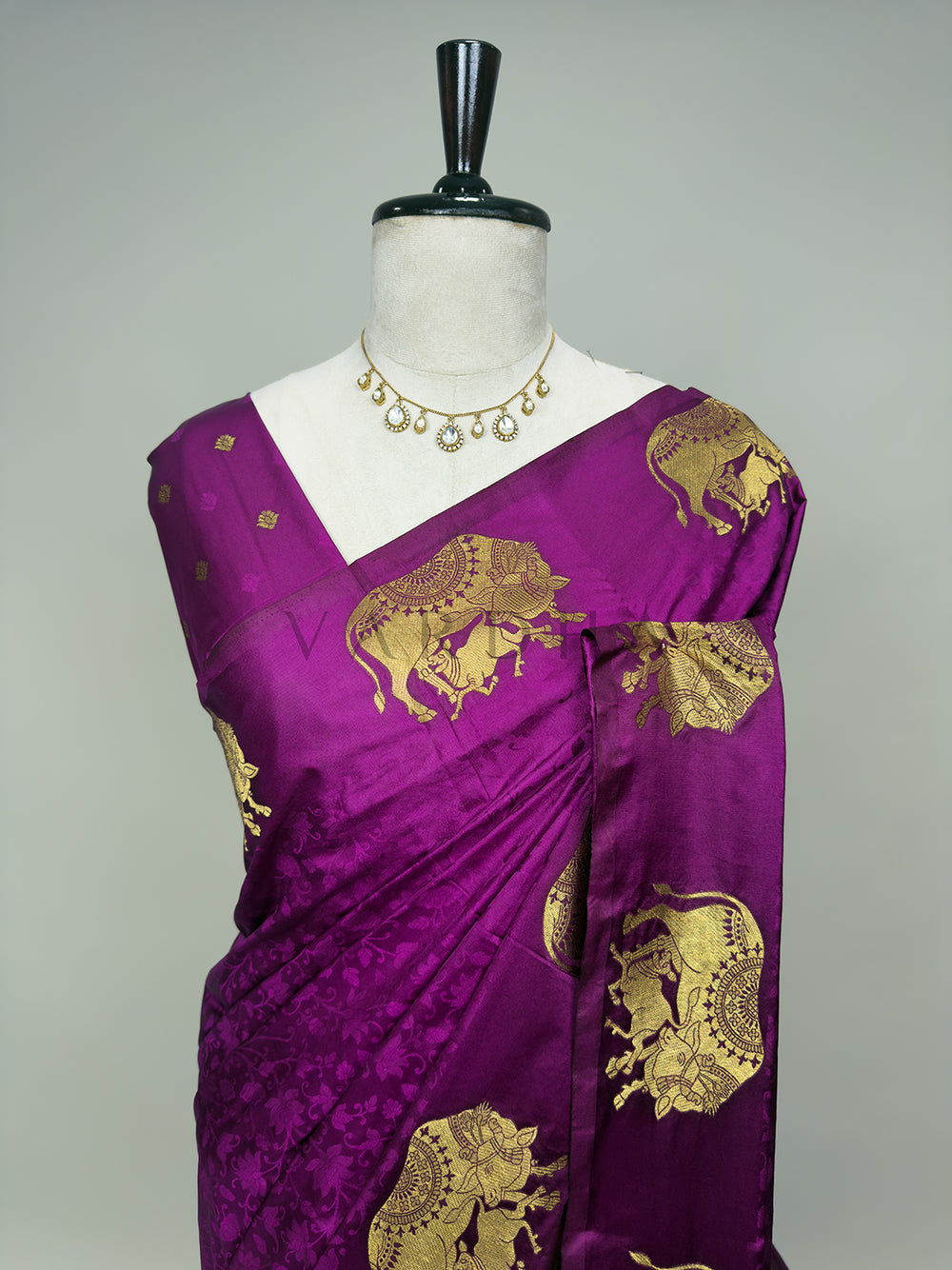 Purple saree with gold elephant patterns on a mannequin against a gray background A detailed half-torso perspective of the draped Royal Magenta Satin Silk Saree on a mannequin, decorated with a classic stone statement necklace. This view highlights the exquisite pattern layout over the chest and pleated shoulder drape, emphasizing the embossed tone-on-tone floral vine designs and the intricately woven gold zari elephant motifs against the glossy magenta base.