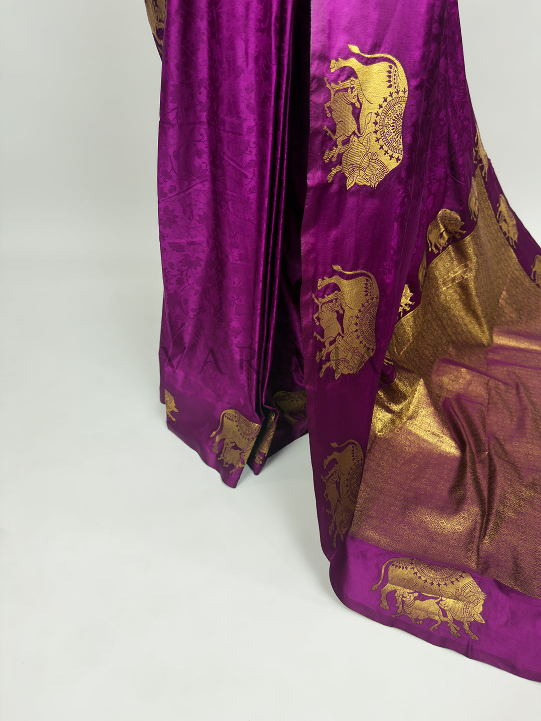 Purple fabric with gold embroidery on a white background A targeted shot focusing on the lower drape and the graceful fall of the Royal Magenta Satin Silk Saree. The image showcases the smooth, lustrous sheen of the premium satin fabric as it forms structured, clean folds. A prominent vertical panel decorated with a repeating sequence of detailed gold zari elephant motifs runs down the side, beautifully accentuating the silhouette.