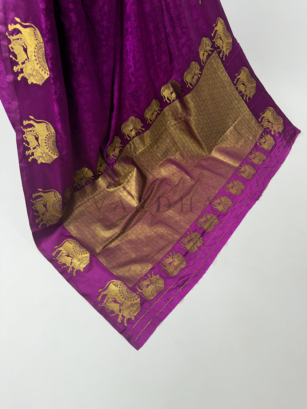 Purple fabric with gold elephant patterns on a white background A high-angle shot showcasing the grand, fully extended pallu of the Royal Magenta Satin Silk Saree spread elegantly across the floor. The center of the palla features a heavy and dense geometric grid pattern woven entirely from shimmering gold zari threads, which is beautifully bordered by a detailed parade of marching elephant motifs, highlighting traditional Banarasi handloom artistry.