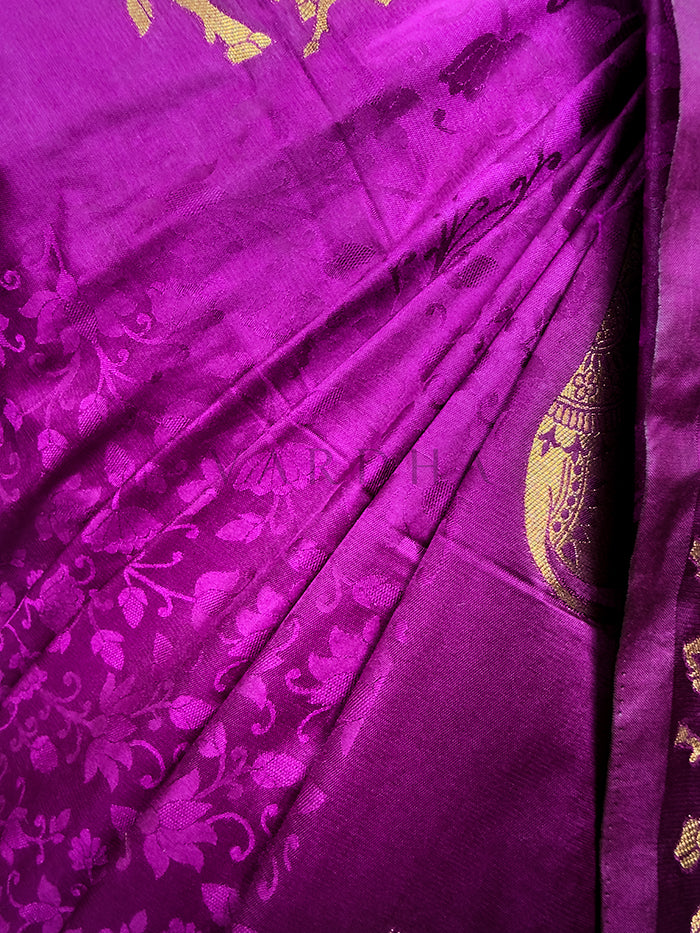 Close-up of a purple fabric with gold floral patterns An extreme close-up view of the Royal Magenta Satin Silk Saree fabric, highlighting its rich texture and immaculate craftsmanship in sharp focus. The shot reveals the complex self-jacquard weaving that creates an embossed pattern of delicate floral vines and leaves across the glossy magenta silk surface, seamlessly paired with the brilliant metallic finish of a detailed gold zari motif.