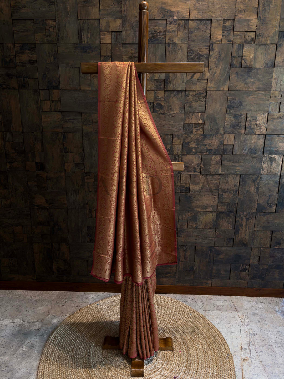 Brown fabric draped over a wooden stand against a textured wall. Rust Red Mysore Silk Saree with intricate golden zari weaving and floral patterns.