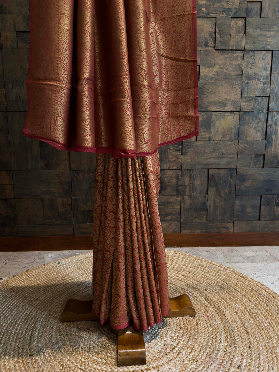 Brown textured fabric draped over a stand against a stone wall. Golden zari woven motifs highlighting traditional Mysore craftsmanship on rust red silk.