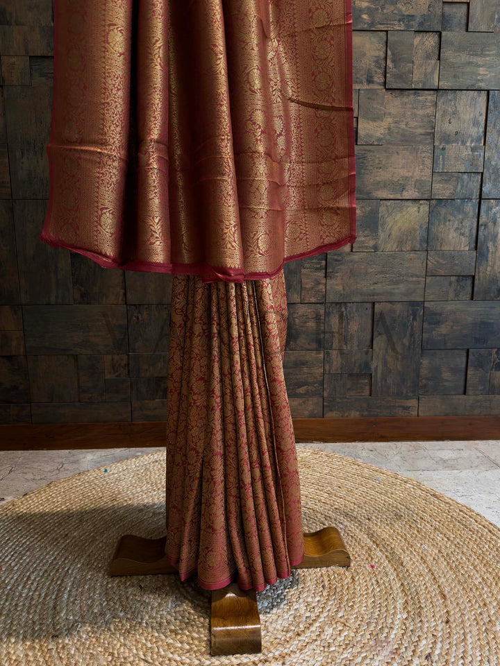 Brown textured fabric draped over a stand against a stone wall. Golden zari woven motifs highlighting traditional Mysore craftsmanship on rust red silk.