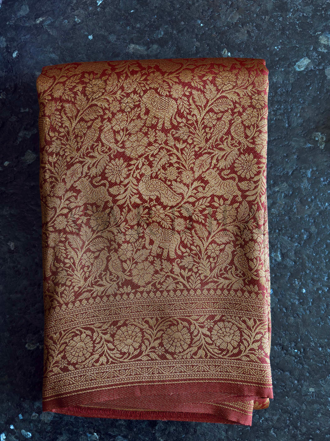 Folded fabric with intricate gold patterns on a dark surface Pallu of rust red Mysore silk saree featuring woven elephant and bird motifs in zari.