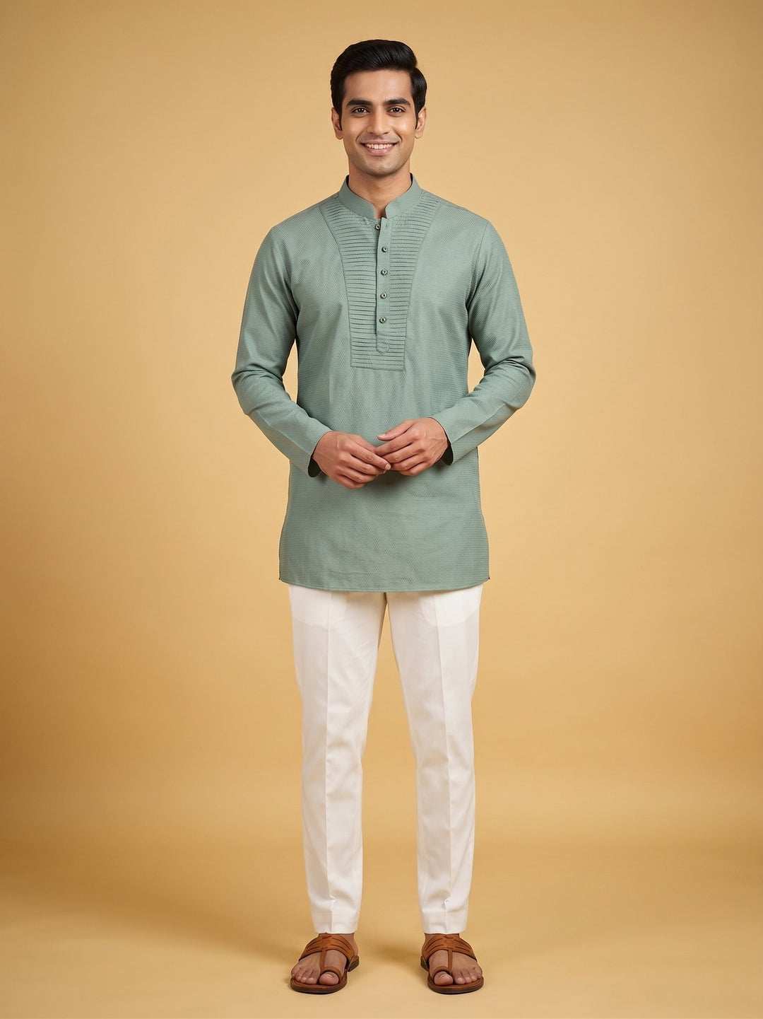 A man modeling a sage green long-sleeve kurta with white trousers and brown sandals, standing against a solid beige studio background.