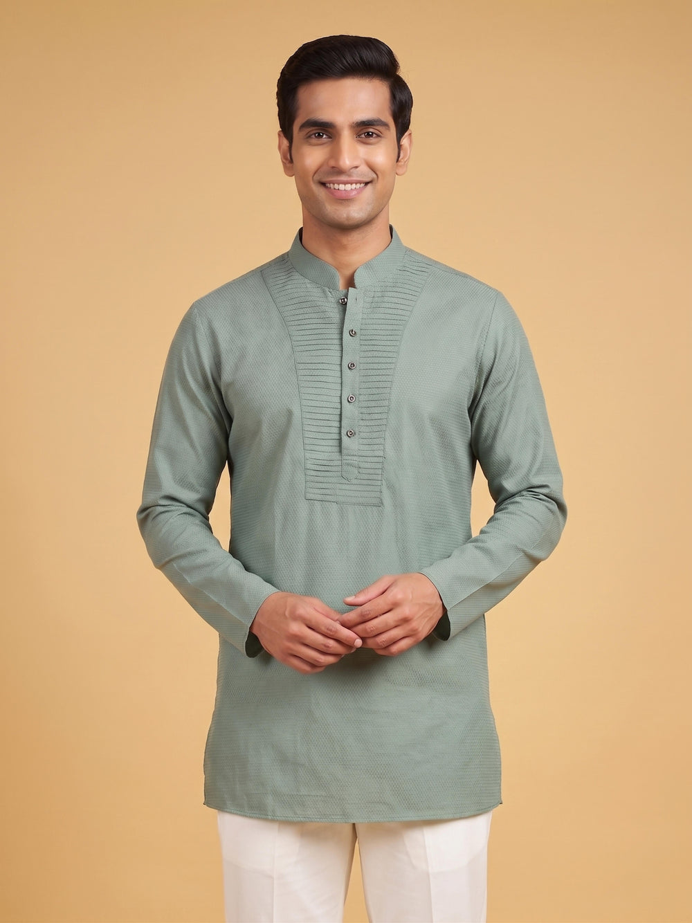 A man wearing a sage green long-sleeve kurta with a mandarin collar and button placket, paired with white trousers, standing against a solid beige background. Medium shot of a man wearing a sage green cotton short kurta with a stylish pintuck yoke and white trousers.