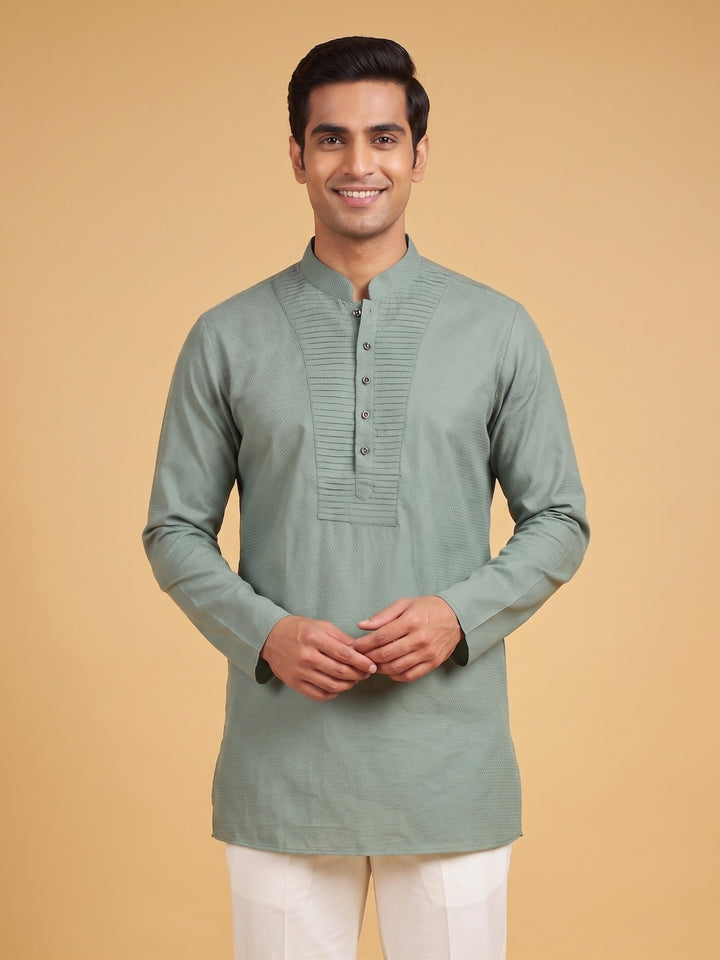 A man wearing a sage green long-sleeve kurta with a mandarin collar and button placket, paired with white trousers, standing against a solid beige background. Medium shot of a man wearing a sage green cotton short kurta with a stylish pintuck yoke and white trousers.