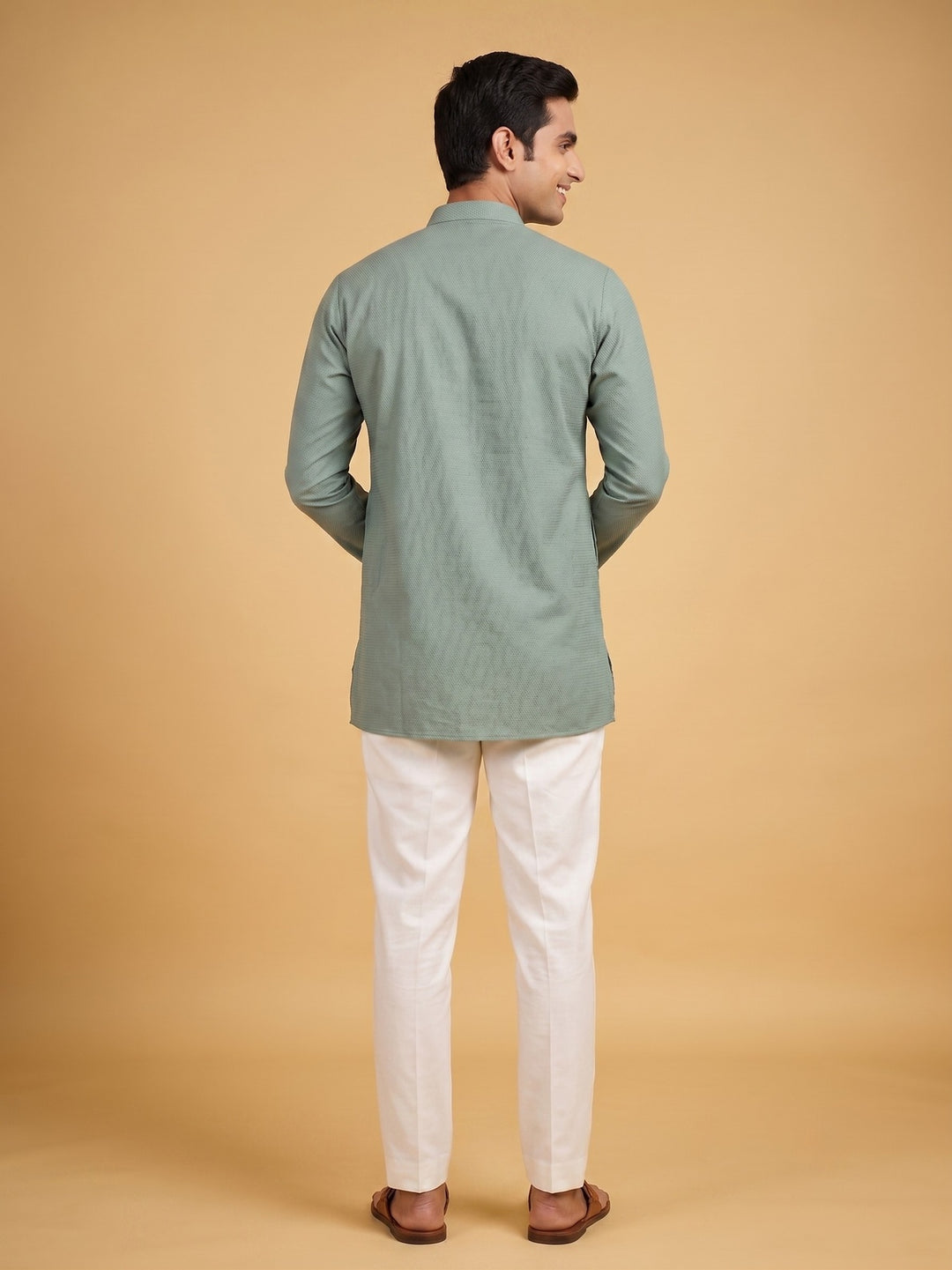 A man shown from the back wearing a sage green long-sleeve kurta and off-white trousers, standing against a solid warm beige background. Back view of a man wearing a sage green cotton short kurta with white trousers.