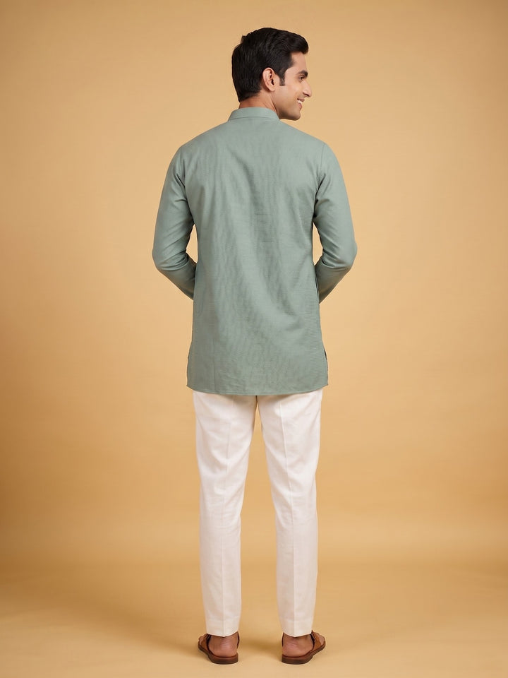A man shown from the back wearing a sage green long-sleeve kurta and off-white trousers, standing against a solid warm beige background. Back view of a man wearing a sage green cotton short kurta with white trousers.