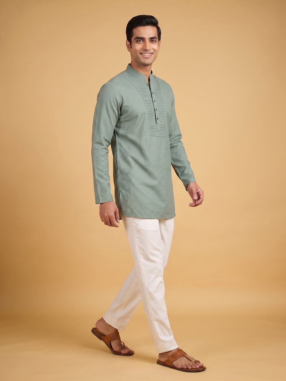 A man modeling a sage green long-sleeve kurta with white trousers and brown sandals, standing against a solid beige studio background. A man walking in a sage green cotton short kurta with a pintuck yoke, side-angle view showcasing fit.