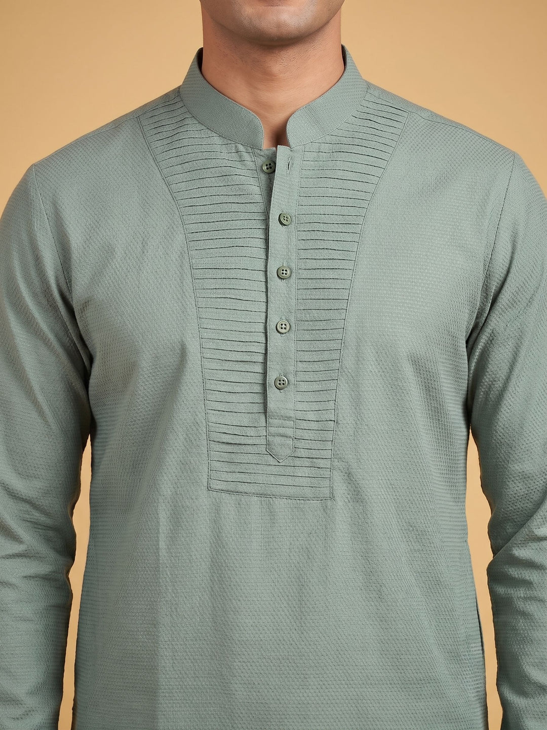 A man wearing a sage green long-sleeve kurta with a textured weave, mandarin collar, and a buttoned pleated chest panel, set against a solid warm beige background. A man standing in a sage green cotton short kurta with a pintuck yoke, relaxed pose.