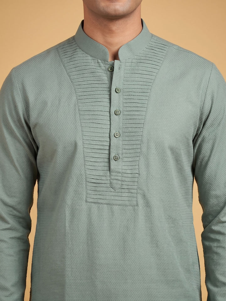 A man wearing a sage green long-sleeve kurta with a textured weave, mandarin collar, and a buttoned pleated chest panel, set against a solid warm beige background. A man standing in a sage green cotton short kurta with a pintuck yoke, relaxed pose.