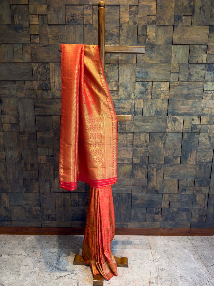 Red saree with gold border on a wooden stand against a textured wall. Sindoori Red Kanjivaram Silk Saree with gold bird motifs and floral zari