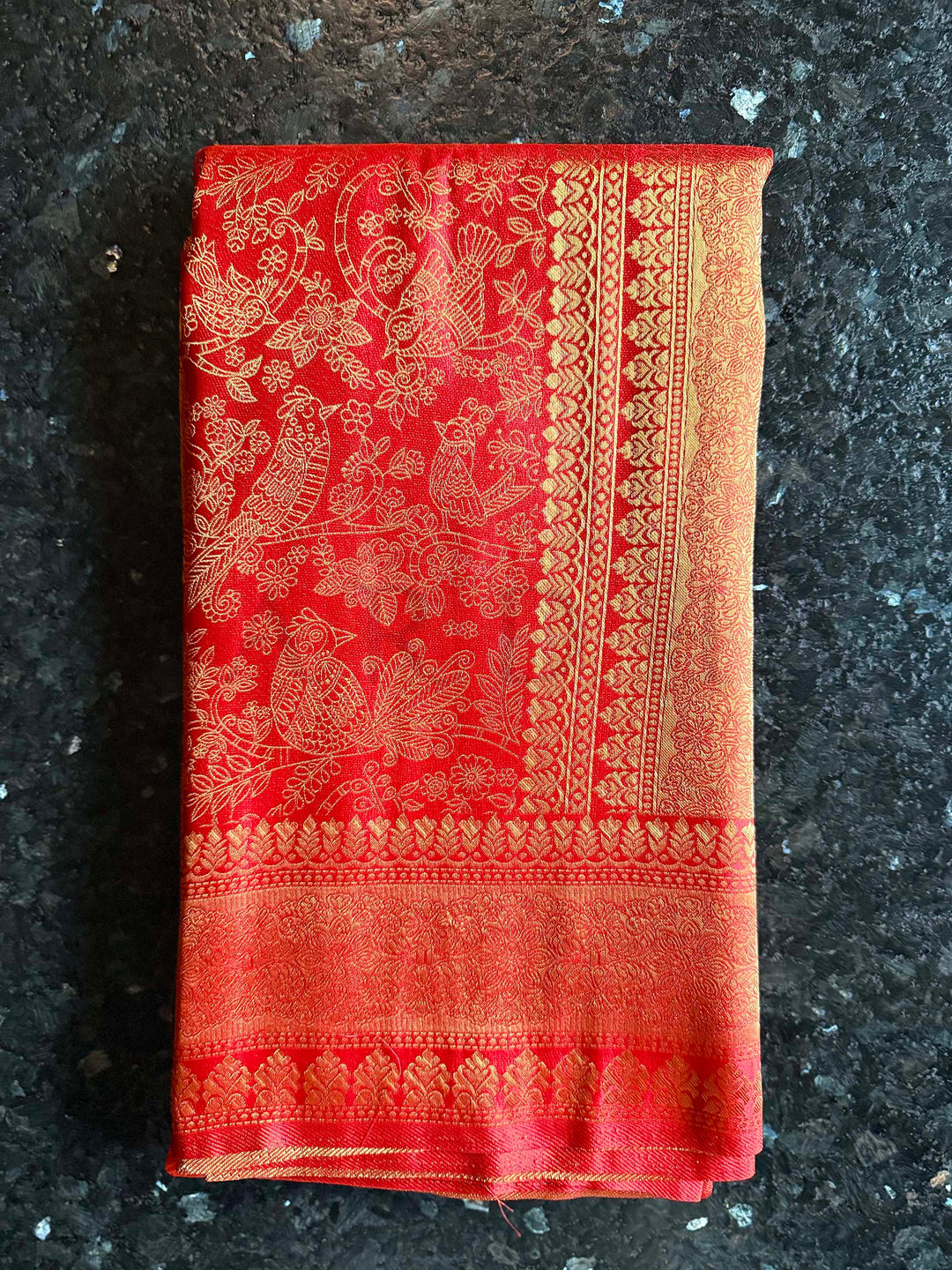 Red fabric with gold embroidery on a dark textured surface, High-detail shot of gold zari craftsmanship on red Kanjivaram border