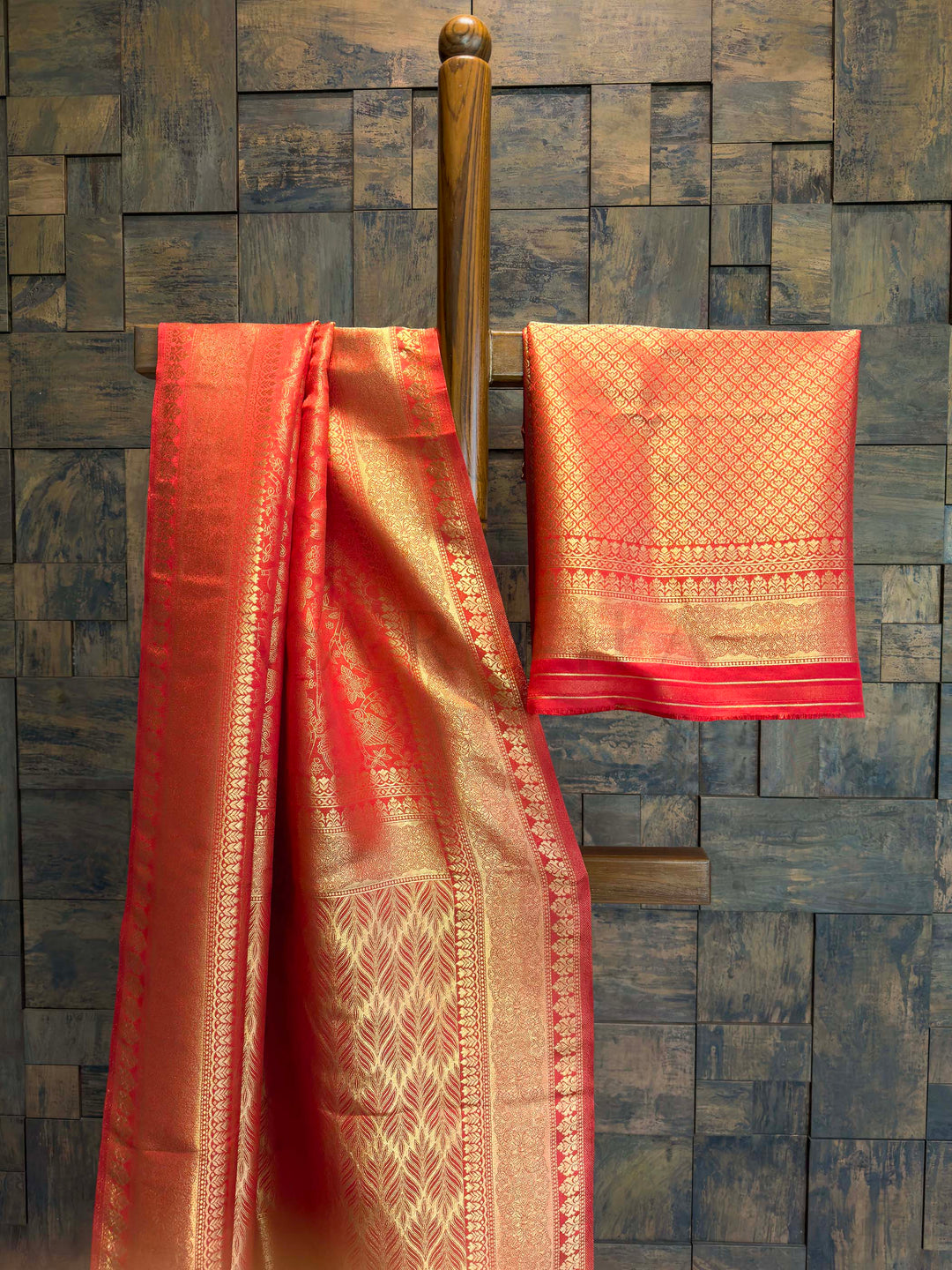 Red and gold saree with a matching blouse draped over a wooden rod against a stone wall. Side drape view showing bird motifs, border detailing, and saree texture