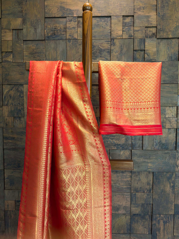 Red and gold saree with a matching blouse draped over a wooden rod against a stone wall. Side drape view showing bird motifs, border detailing, and saree texture