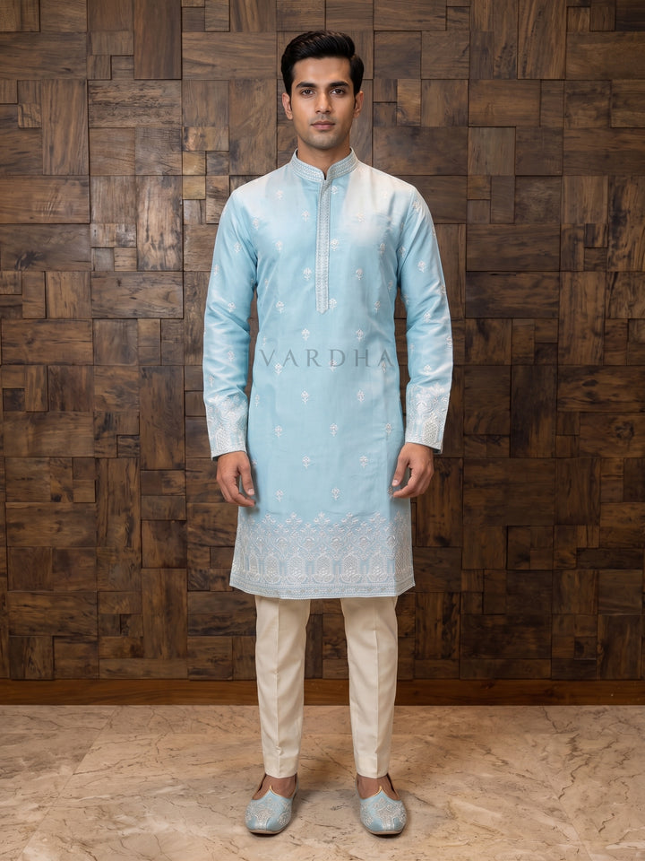 Man wearing a light blue kurta with white embroidery against a wooden paneled wall. Art Silk, the kurta offers a luxurious sheen and a flawless drape. The body is delicately adorned with scattered white floral buttas, leading down to a masterpiece of a hemline. The heavy border embroidery on the daman (hem) and sleeve cuffs features intricate architectural and floral motifs