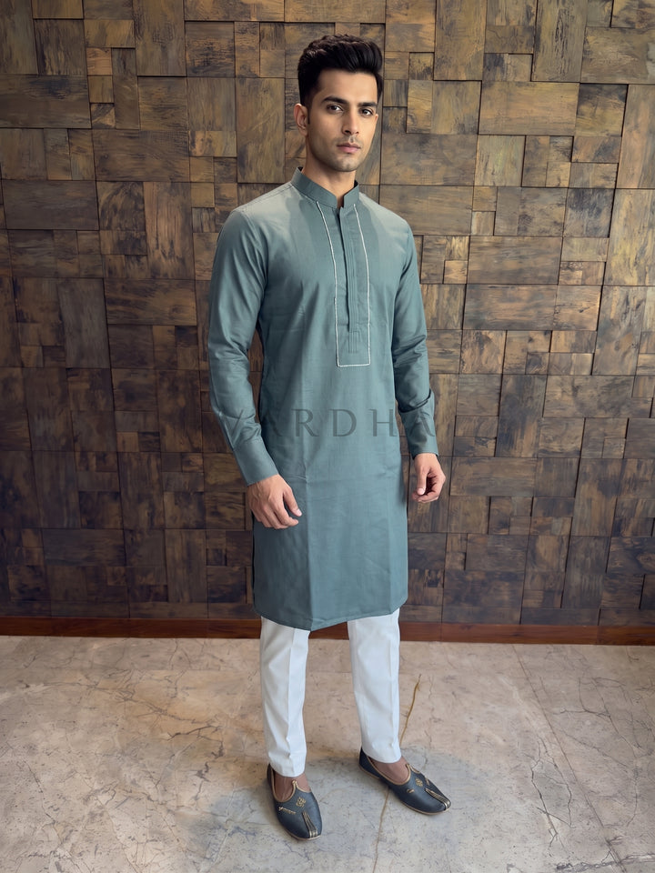 Man wearing a green kurta with white pants against a wooden paneled wall. A knee-length Slate Green cotton kurta for men featuring a structured chest design with vertical pintucks and a formal Mandarin collar.