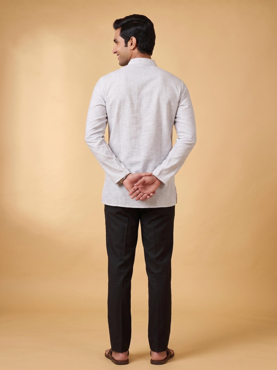 A man shown from the back wearing a light gray long-sleeve kurta and black trousers, standing against a solid beige studio background. Back view of a man showcasing the fit and length of a steel grey cotton short kurta paired with black trousers.