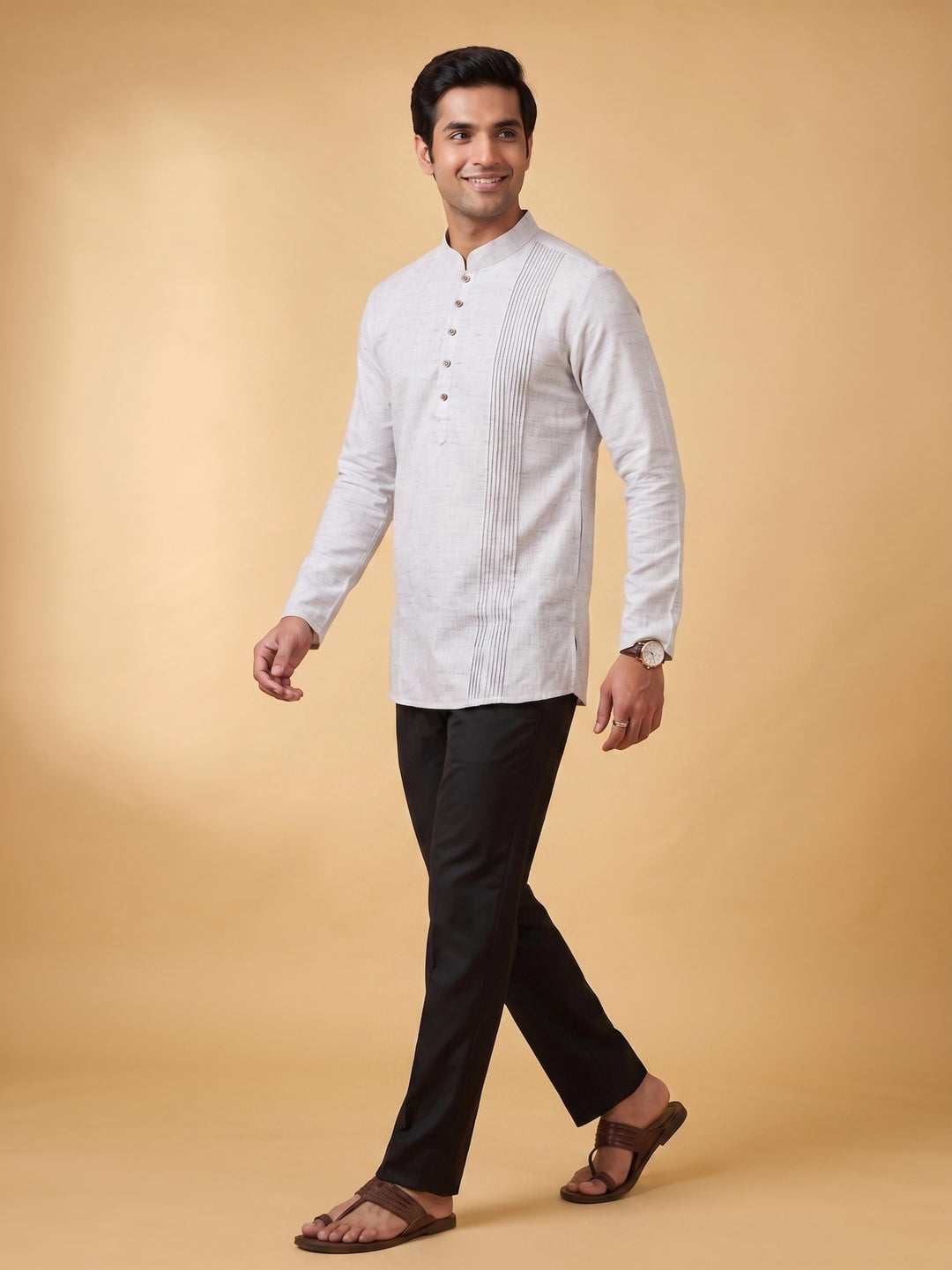A man modeling a white long-sleeve kurta with vertical pleats and a band collar, paired with black trousers and brown sandals, against a solid beige studio background. A man walking in a steel grey cotton short kurta with a structured pintuck yoke, side-angle view.