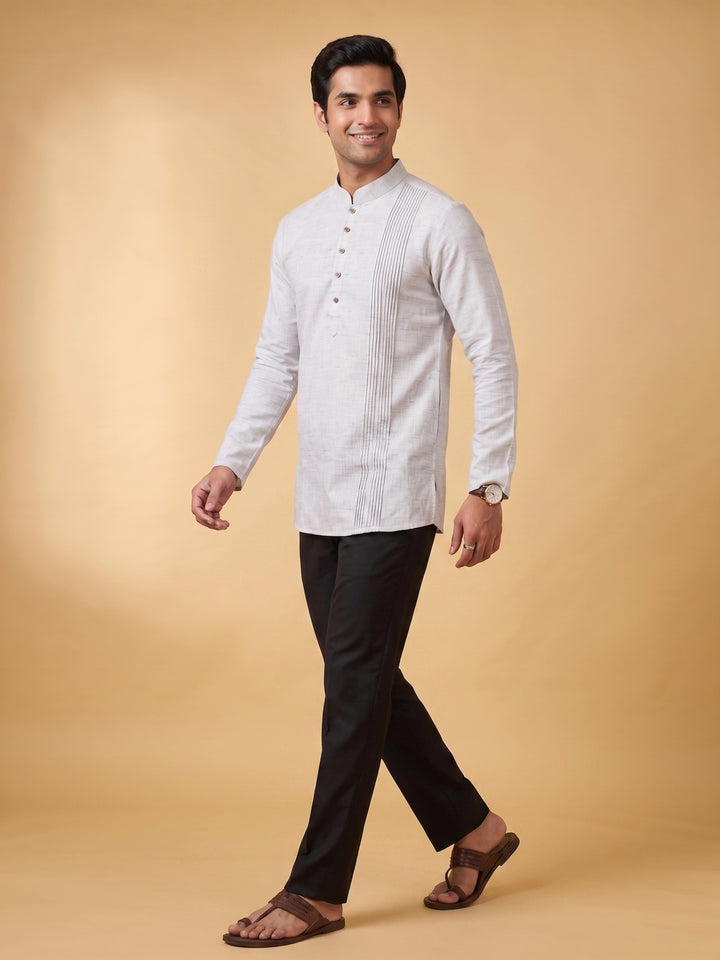 A man modeling a white long-sleeve kurta with vertical pleats and a band collar, paired with black trousers and brown sandals, against a solid beige studio background. A man walking in a steel grey cotton short kurta with a structured pintuck yoke, side-angle view.