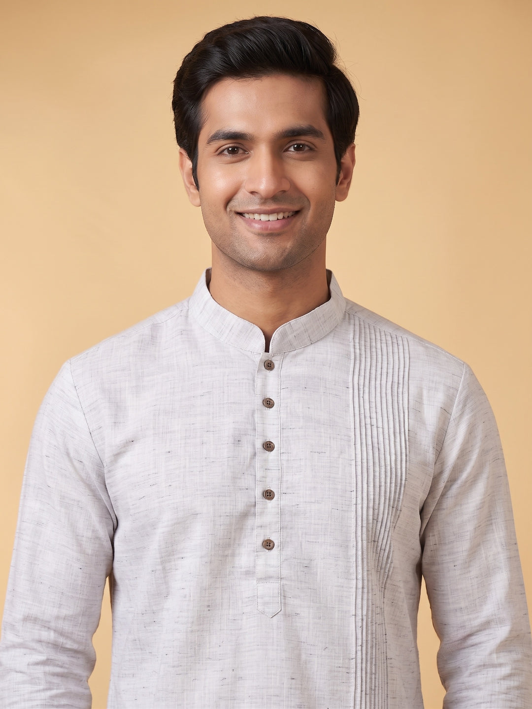 A man wearing a light gray textured kurta with a mandarin collar and button placket, standing against a solid beige background. Detailed medium shot of a man wearing a steel grey cotton short kurta, highlighting the structured pintuck yoke design.