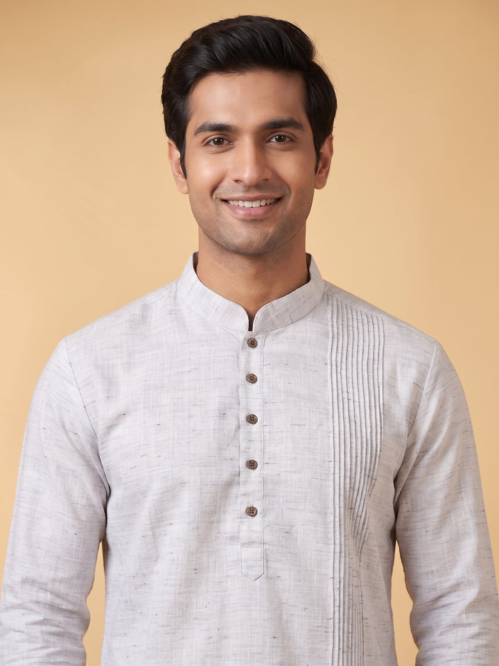 A man wearing a light gray textured kurta with a mandarin collar and button placket, standing against a solid beige background. Detailed medium shot of a man wearing a steel grey cotton short kurta, highlighting the structured pintuck yoke design.