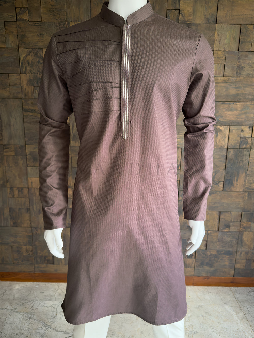 Brown kurta on a mannequin against a wooden paneled wall Close-up of the dusty mauve brown men's festive kurta showcasing the fine micro-texture, unique chest pleating, and a concealed placket with contrast triple-stitch detailing.