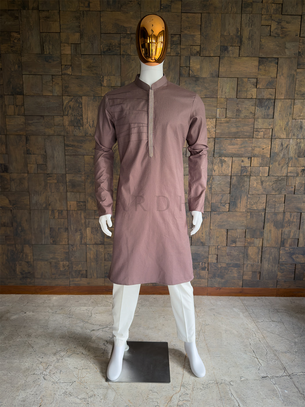 Mannequin wearing a Brown kurta with a gold mask against a wooden paneled wall. Front view of a man wearing a House of Vardha taupe brown textured cotton kurta featuring asymmetrical pintuck pleats. Perfect men's ethnic wear for festive parties and traditional smart-casual events.