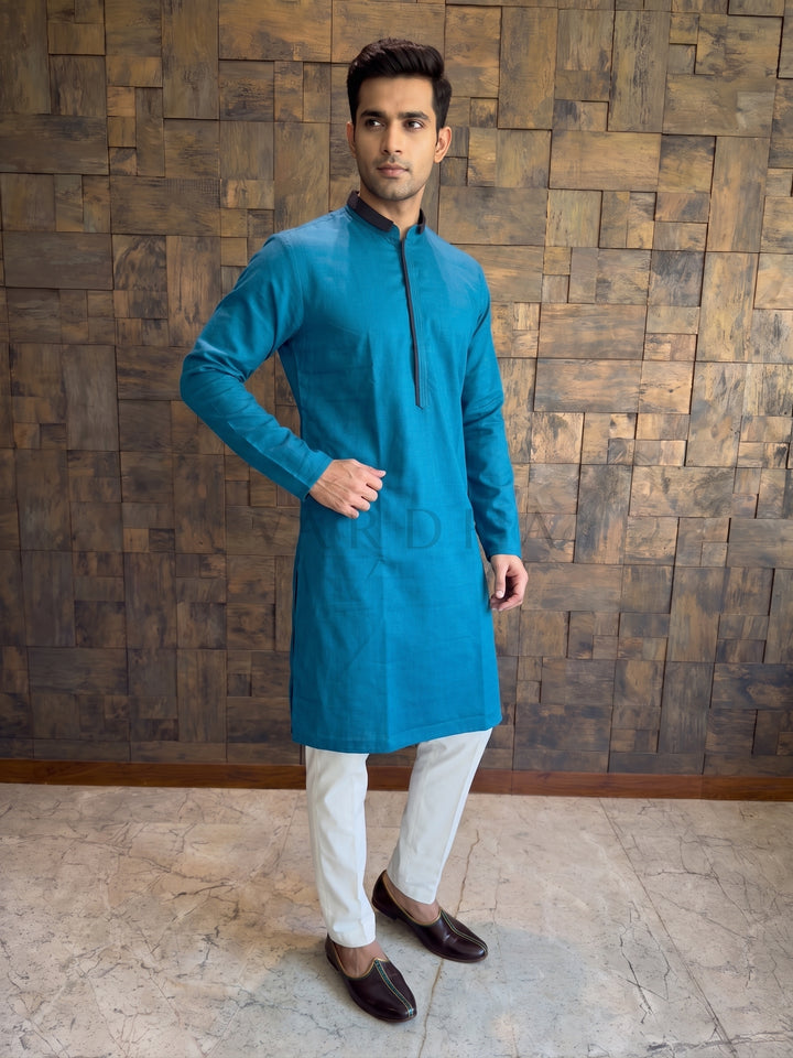 Man wearing a blue kurta with white pants against a wooden paneled wall. Vibrant Teal Blue cotton kurta for men featuring a sharp dark contrast Mandarin collar and a sleek concealed placket framed with matching dark piping.