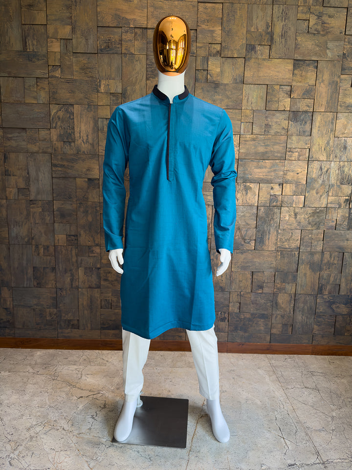 Mannequin wearing a blue kurta against a wooden paneled wall. Modern minimalist ethnic wear: A knee-length Teal Blue kurta with sharp, uninterrupted lines and a highly structured silhouette for a bold statement.