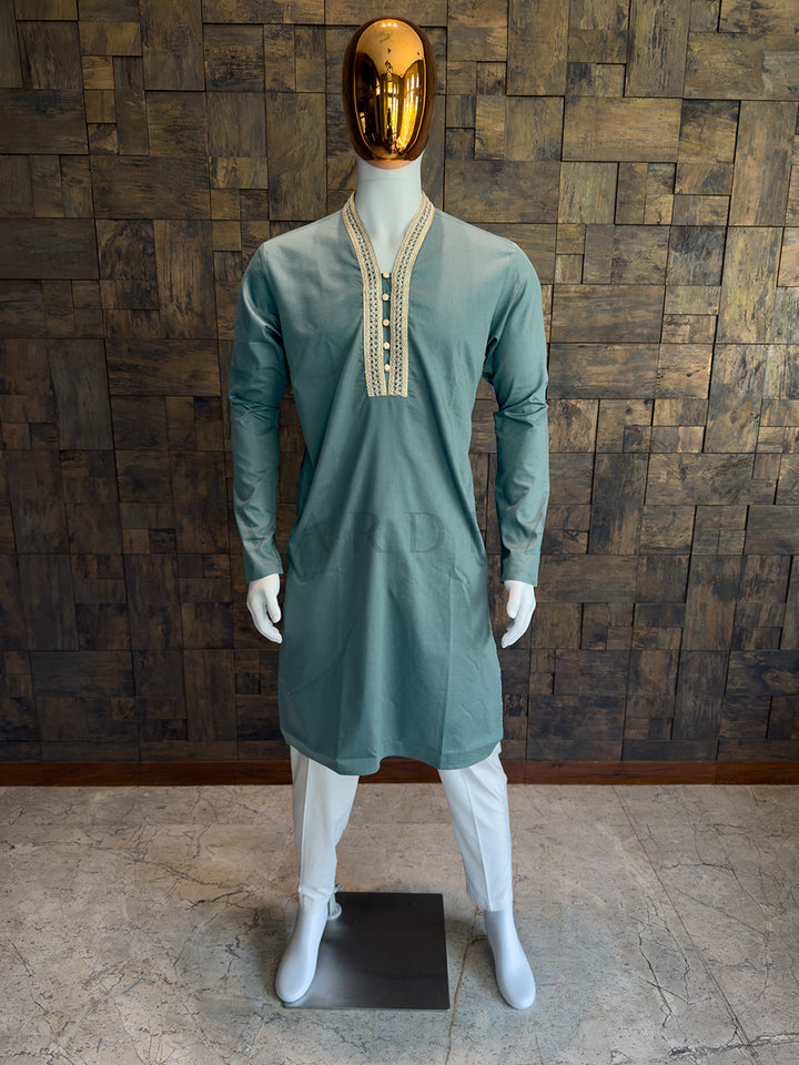 Mannequin wearing a teal blue kurta with white pants against a wooden paneled wall. A knee-length Teal Blue cotton kurta for men, blending a contemporary collarless silhouette with traditional gold woven craftsmanship.