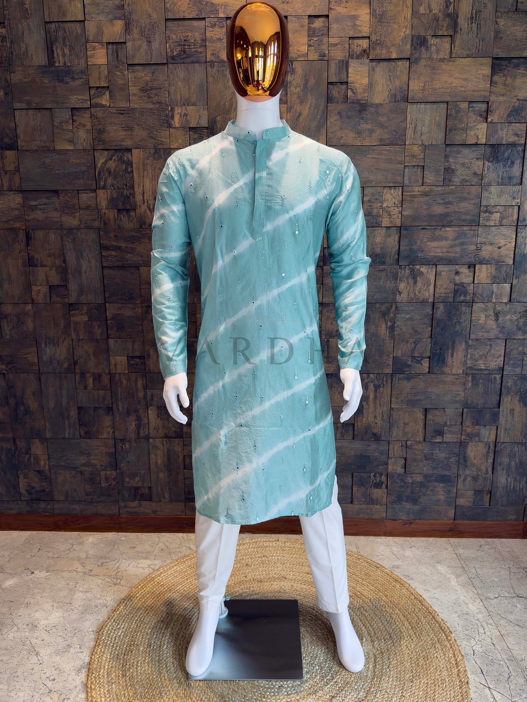 Mannequin wearing a light green kurta with white patterns against a wooden paneled wall. Mint teal viscose men’s kurta with mandarin collar, concealed pintuck placket and diagonal tie-dye bands accented by scattered sequins, styled with white churidar.