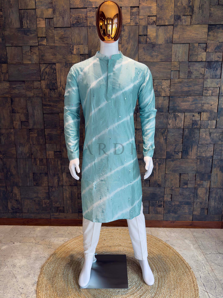 Mannequin wearing a light green kurta with white patterns against a wooden paneled wall. Mint teal viscose men’s kurta with mandarin collar, concealed pintuck placket and diagonal tie-dye bands accented by scattered sequins, styled with white churidar.