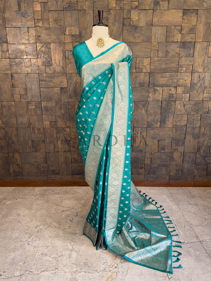 Teal saree with white patterns on a mannequin against a wooden paneled wall. Full front display of a teal green satin silk saree featuring woven zari paisley bootis draped gracefully on a mannequin. Elegant party wear teal satin silk saree highlighting its smooth drape, lustrous shine, and exceptional zari craftsmanship.