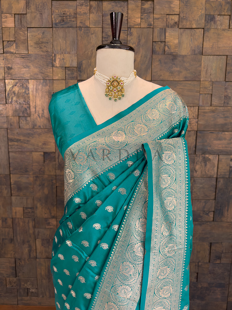 Teal saree with silver patterns on a mannequin against a wooden paneled wall. Close-up of a bright teal woven satin ethnic saree styled on a mannequin display, paired with a traditional pearl choker necklace. Festive ethnic wear sea green zari woven saree sweeping the floor, showcasing the heavily brocaded pallu and traditional patterns.