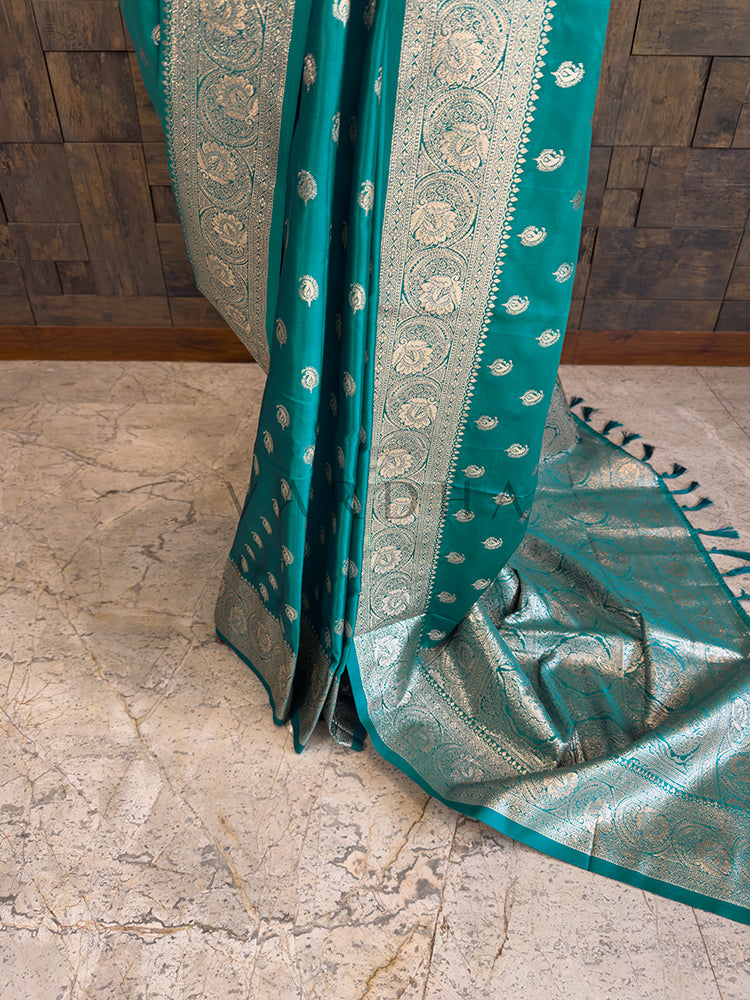 Teal saree with silver border on a stone floor Lower pleats and fluid drape of a glossy teal Banarasi-style satin silk saree showcasing the broad circular floral border.