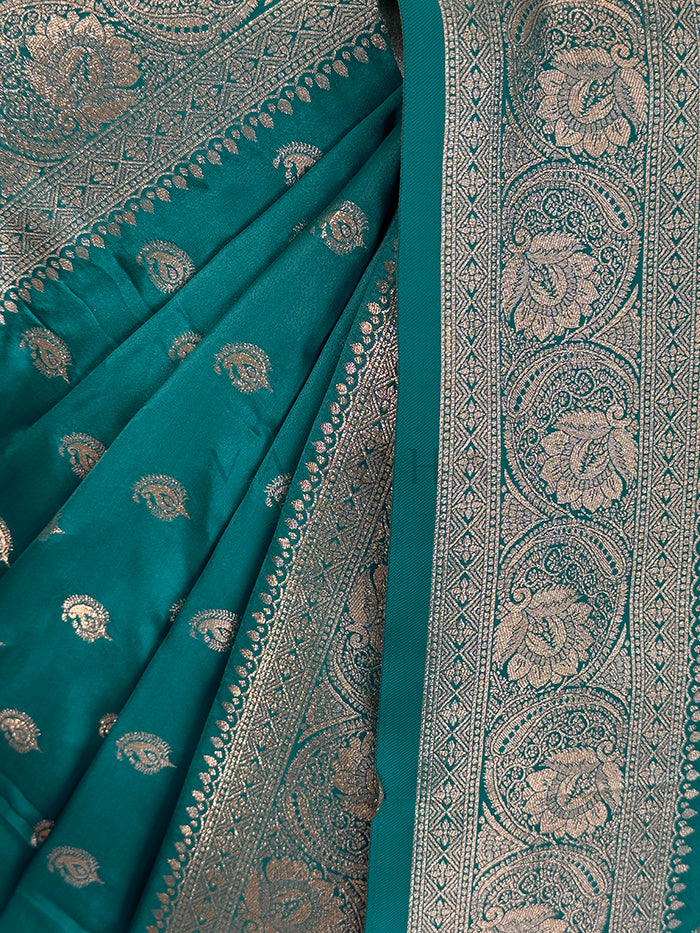 Teal saree with intricate silver patterns on a plain background Close-up texture shot highlighting the woven paisley bootis and dense traditional zari border on a teal green satin fabric.