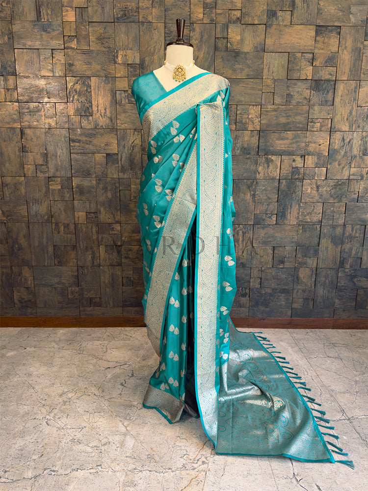 Teal saree with white patterns on a mannequin against a wooden paneled wall. Full front display of a teal green satin silk saree featuring golden zari bootis draped gracefully on a mannequin.