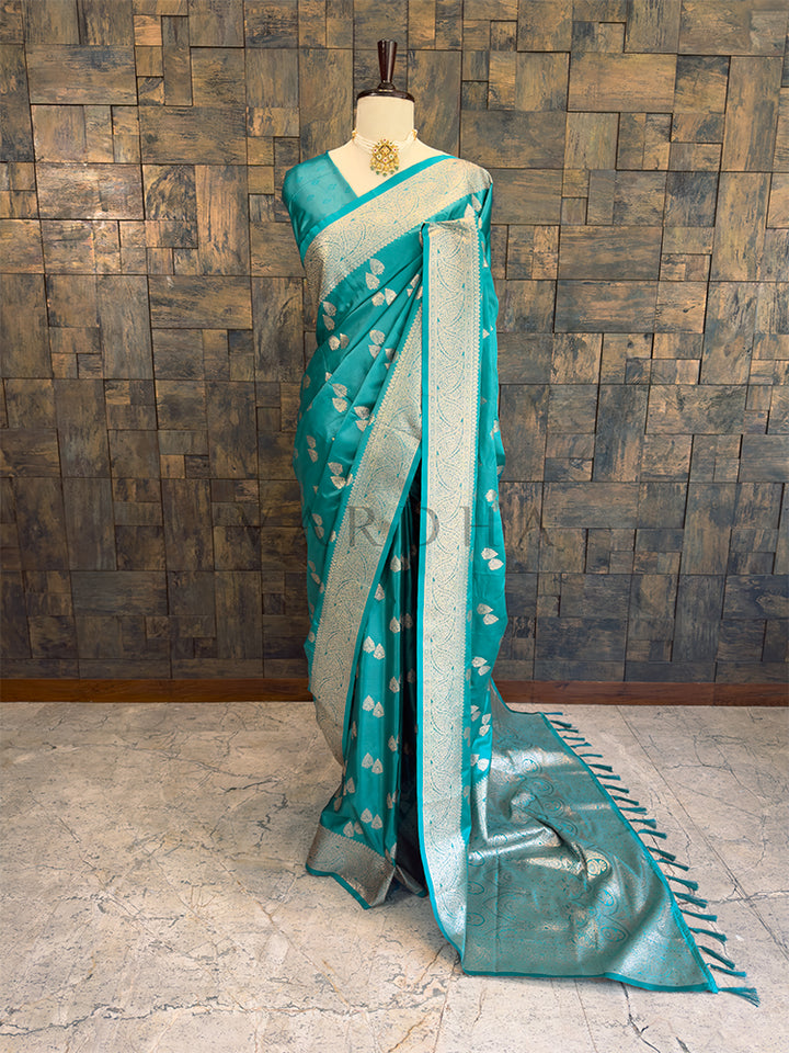Teal saree with white patterns on a mannequin against a wooden paneled wall. Full front display of a teal green satin silk saree featuring golden zari bootis draped gracefully on a mannequin.