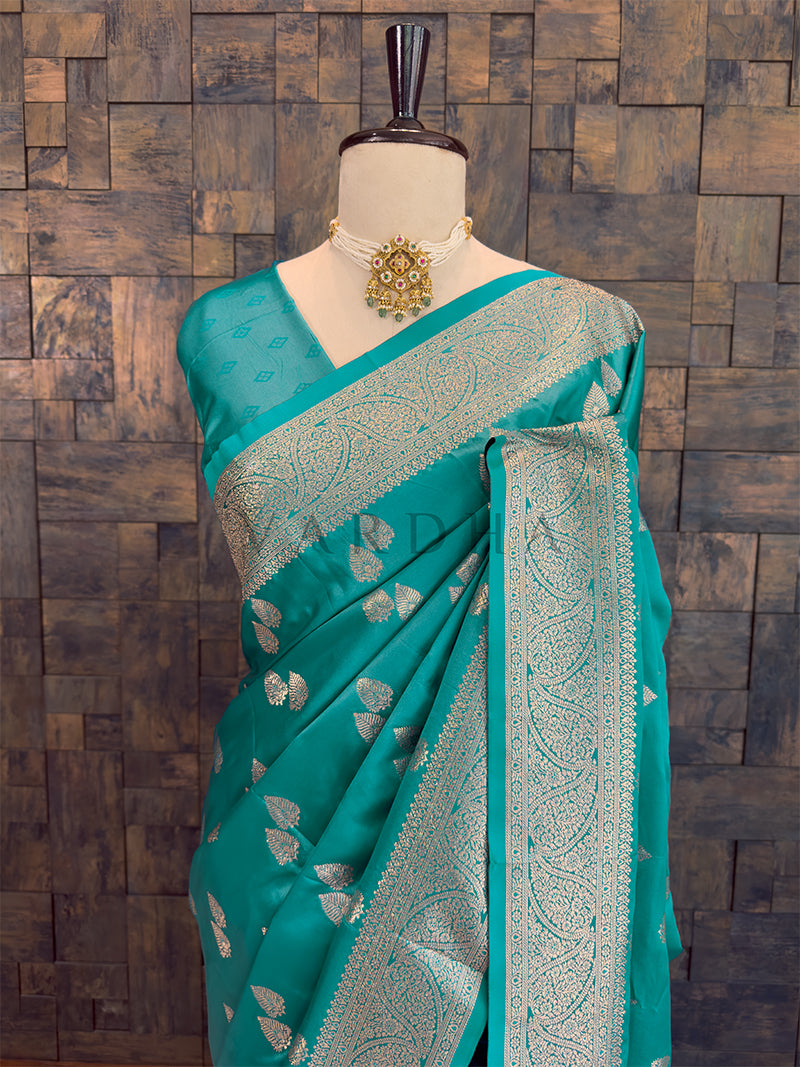 Teal saree with silver patterns on a mannequin against a wooden paneled wall. Close-up of a teal woven satin ethnic saree styled on a mannequin display, paired with a traditional choker necklace.