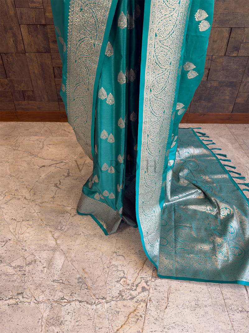 Teal fabric with silver patterns on a stone floor Detailed floor view of the rich, heavy zari woven pallu with matching teal tassels on the luxurious satin saree. Lower pleats and fluid drape of a glossy peacock green Banarasi-style satin silk saree.