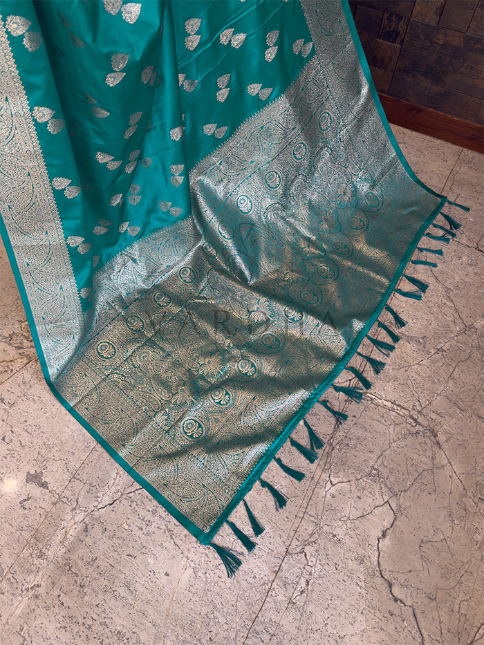Teal and silver patterned fabric with tassels on a stone surface Close-up texture shot highlighting the woven leaf bootis and dense floral zari border on a teal green satin fabric. Elegant party wear teal green satin silk saree highlighting its smooth drape, lustrous shine, and intricate zari craftsmanship.